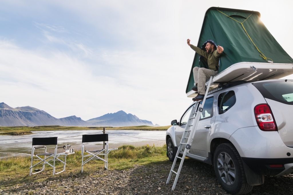 Young bearded man sitting and stretching in a roof top tent mounted on offroad car in Hofn camping site